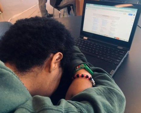 A student in a green sweatshirt puts their head on a desk in front of an open computer.