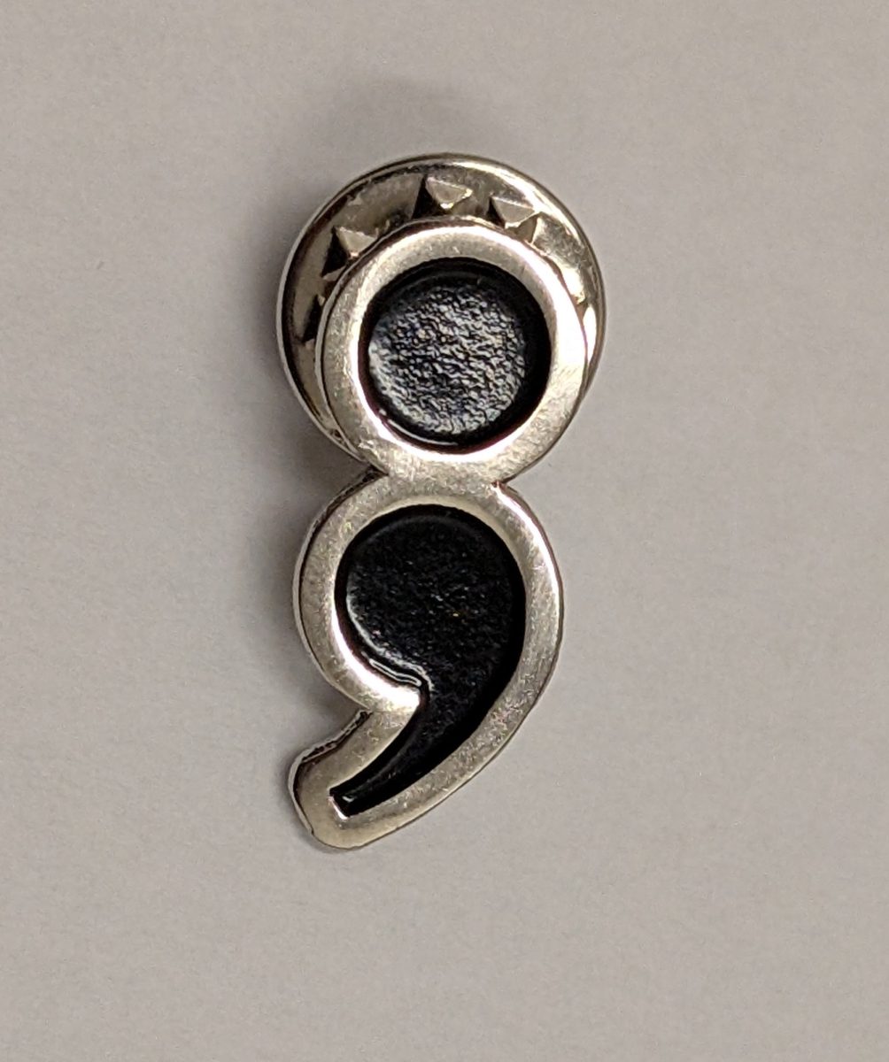 The semi-colon, like the one on this pin, has become a symbol of solidarity around mental health issues, and specifically for suicide awareness.